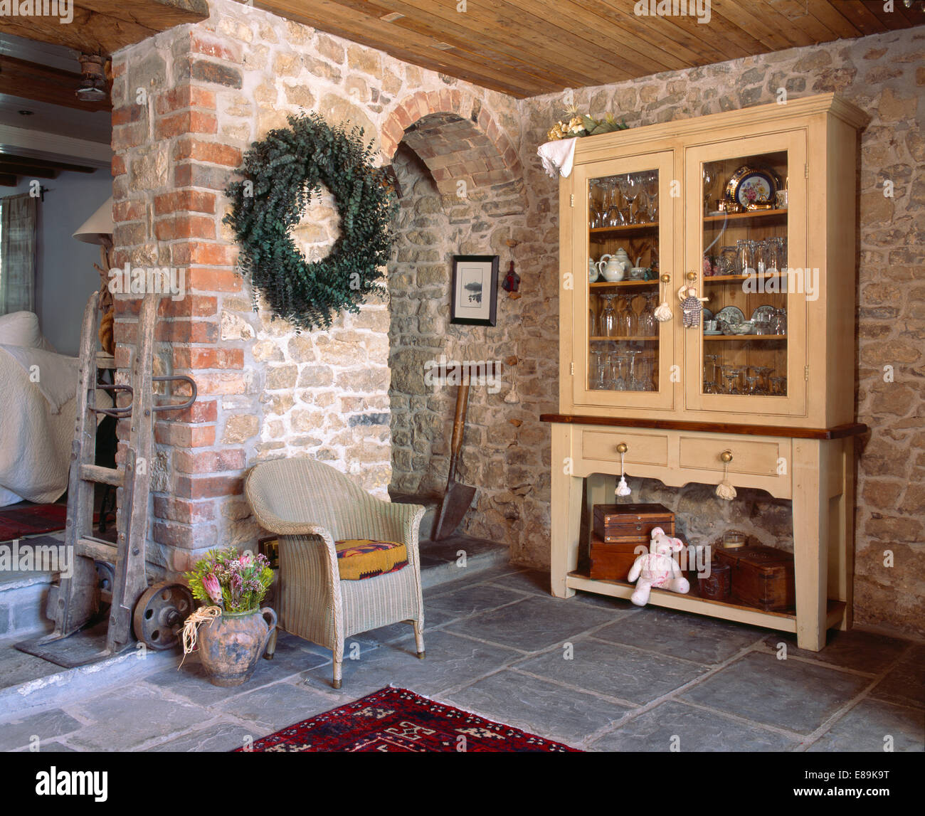 Großer Kranz über Lloyd Loom Stuhl im Wohnzimmer mit rustikalen Steinmauern und französischen Glasfront Kabinett Stockfoto