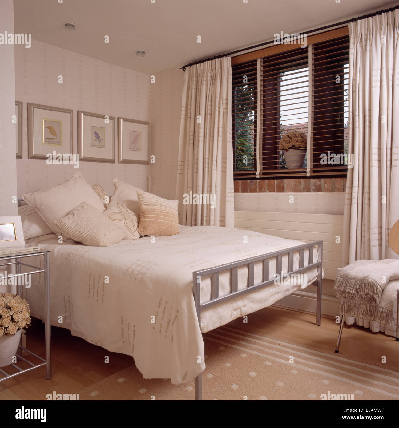 Sahne-Kissen und decken auf einfache Metall Bett in modernen Schlafzimmer mit cremefarbenen Vorhängen und Jalousie am Fenster Stockfoto