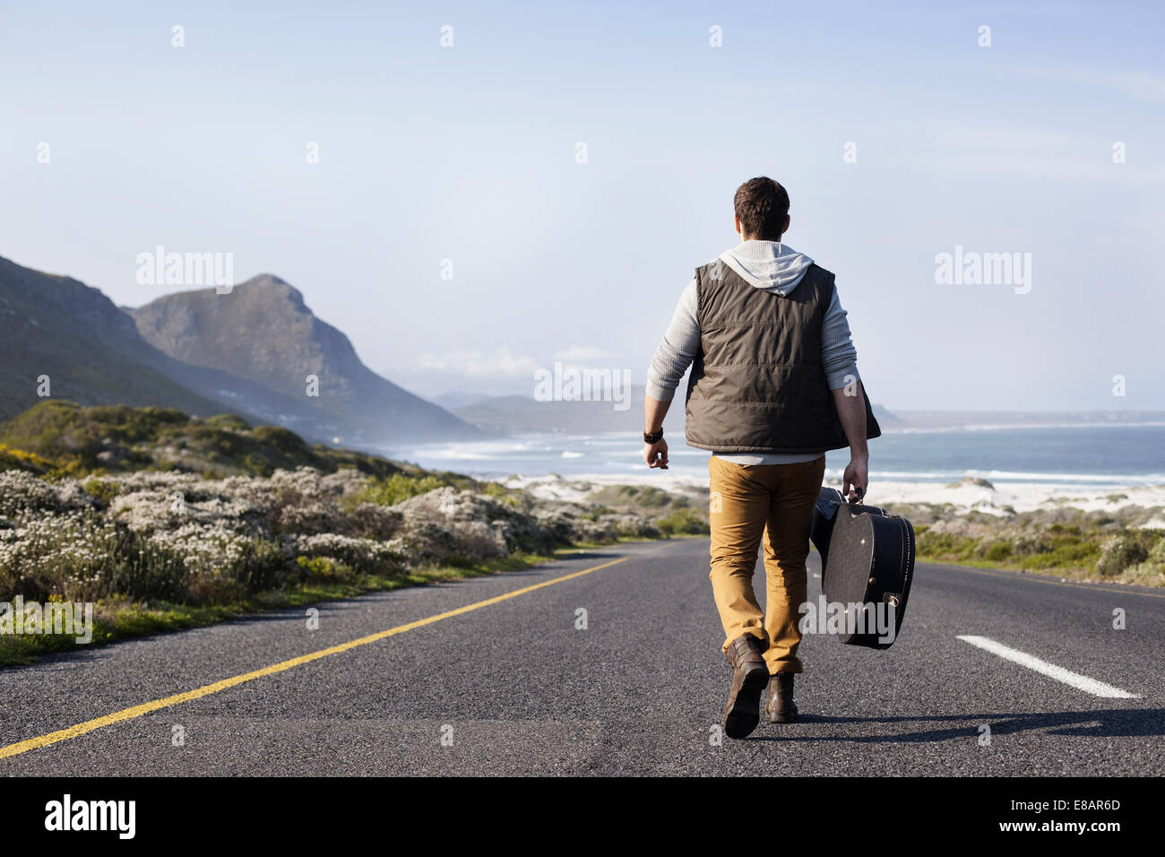 Rückansicht des jungen Mannes mit Gitarrenkoffer zu Fuß auf der Küstenstraße, Cape Town, Western Cape, Südafrika Stockfoto