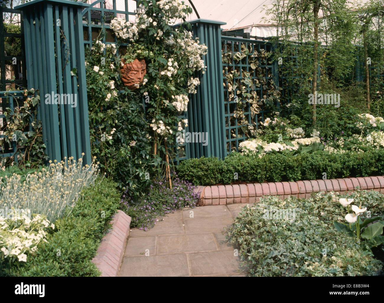 Weiße Rosen auf Zaun mit Säulen im Stadtgarten mit duftenden Pflanzen wachsen in Grenze mit gemauerten Einfassung neben Weg Stockfoto