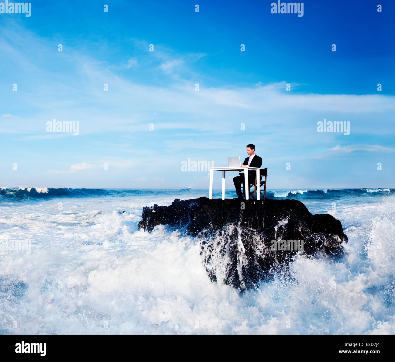 Einsamer Geschäftsmann auf einem abgelegenen Felsen mit sprudelnden Wellen um ihn herum arbeiten. Stockfoto