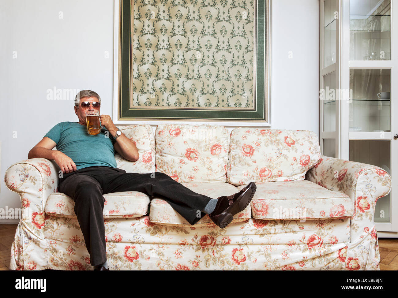 Ein älterer Mann breitet sich beim Entspannen ein Kreuz ein Sofa. Er ist ein Bier trinken und direkt in die Kamera schauen. Stockfoto