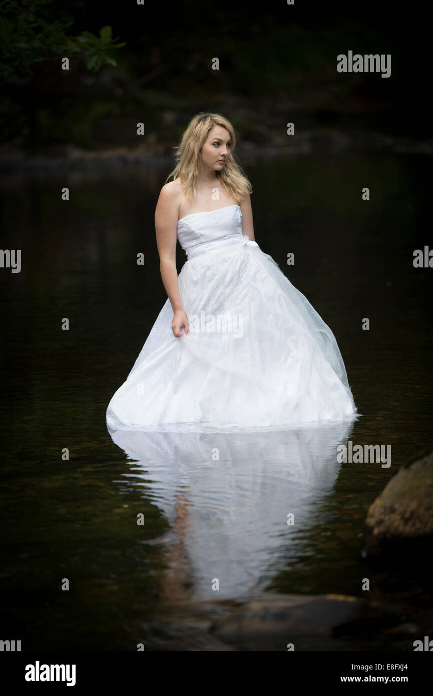 "Trash The Dress": eine junge blonde Frau Mädchen Modell Braut tragen weiße Ballkleid Brautkleid in langsam fließenden Fluss stehend Stockfoto