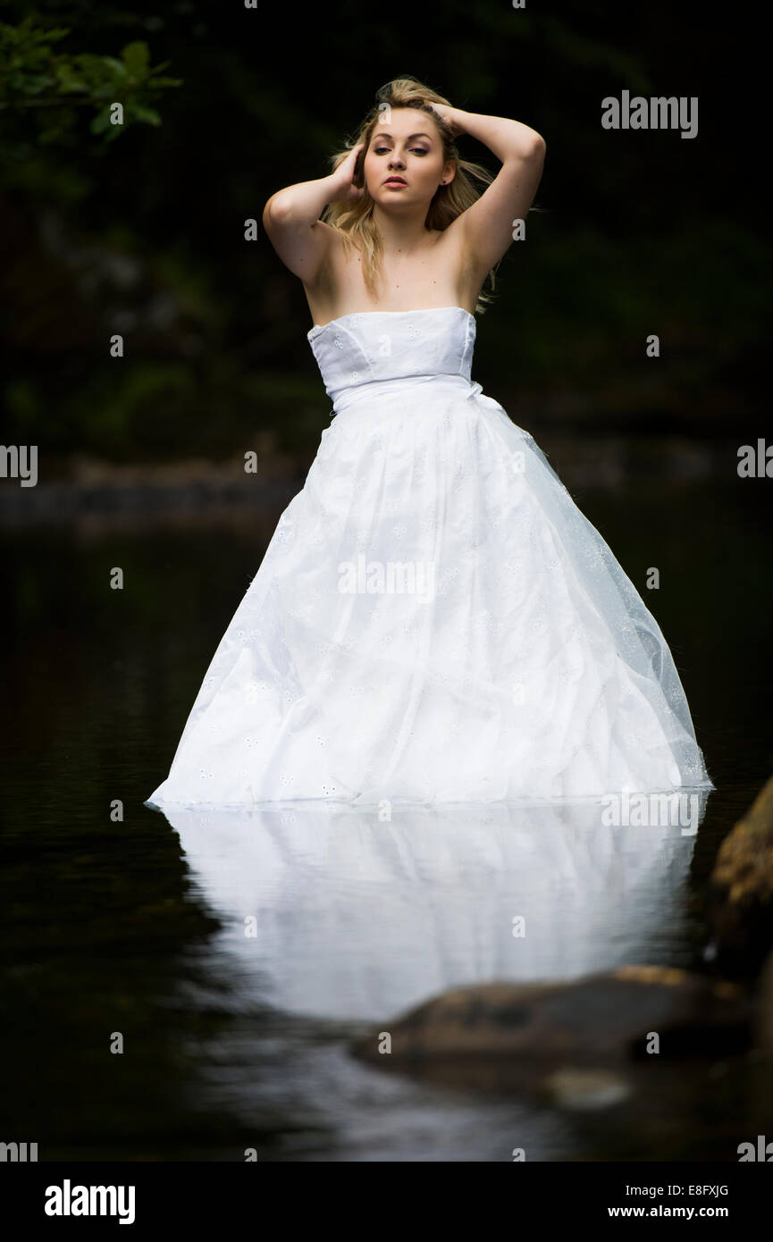 "Trash The Dress": eine junge blonde Frau Mädchen Modell Braut tragen weiße Ballkleid Brautkleid in langsam fließenden Fluss stehend Stockfoto