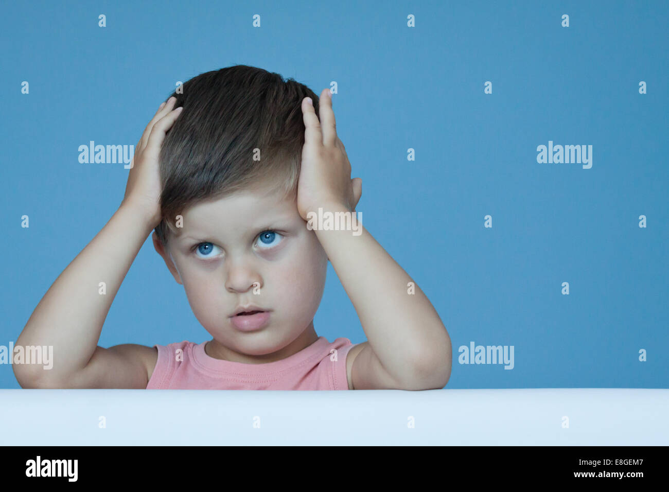 Drei Jahre alte junge hält Hand auf Gesicht ausdrückliche Gefühle wie Verlegenheit Nachdenklichkeit über blaue Wand Hintergrund posiert Stockfoto