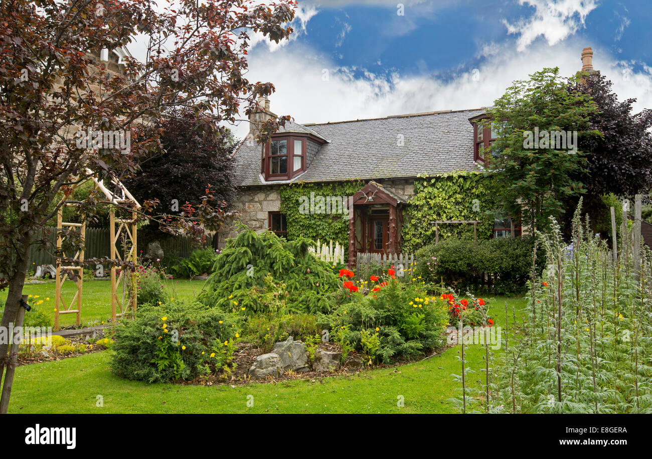 Stein eingemauert Cottage mit gepflegten Garten mit bunten Frühlingsblumen, Bäume, smaragdgrünen Rasen, Torbogen über Pfad, unter blauem Himmel Stockfoto
