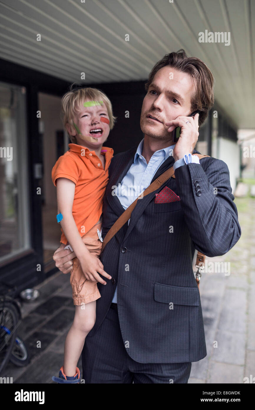 Geschäftsmann mit Handy während des Tragens weinenden Sohn vor Haus Stockfoto