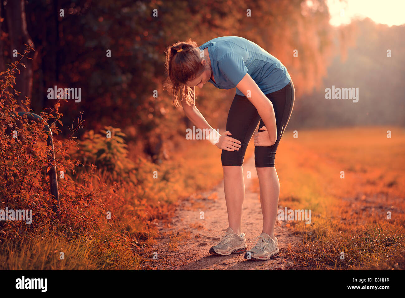 Junge sportliche Frau, die eine Pause vom Training stehen ruht ihr die Hände auf den Knien auf einem ländlichen Weg durch üppige Ackerland Stockfoto