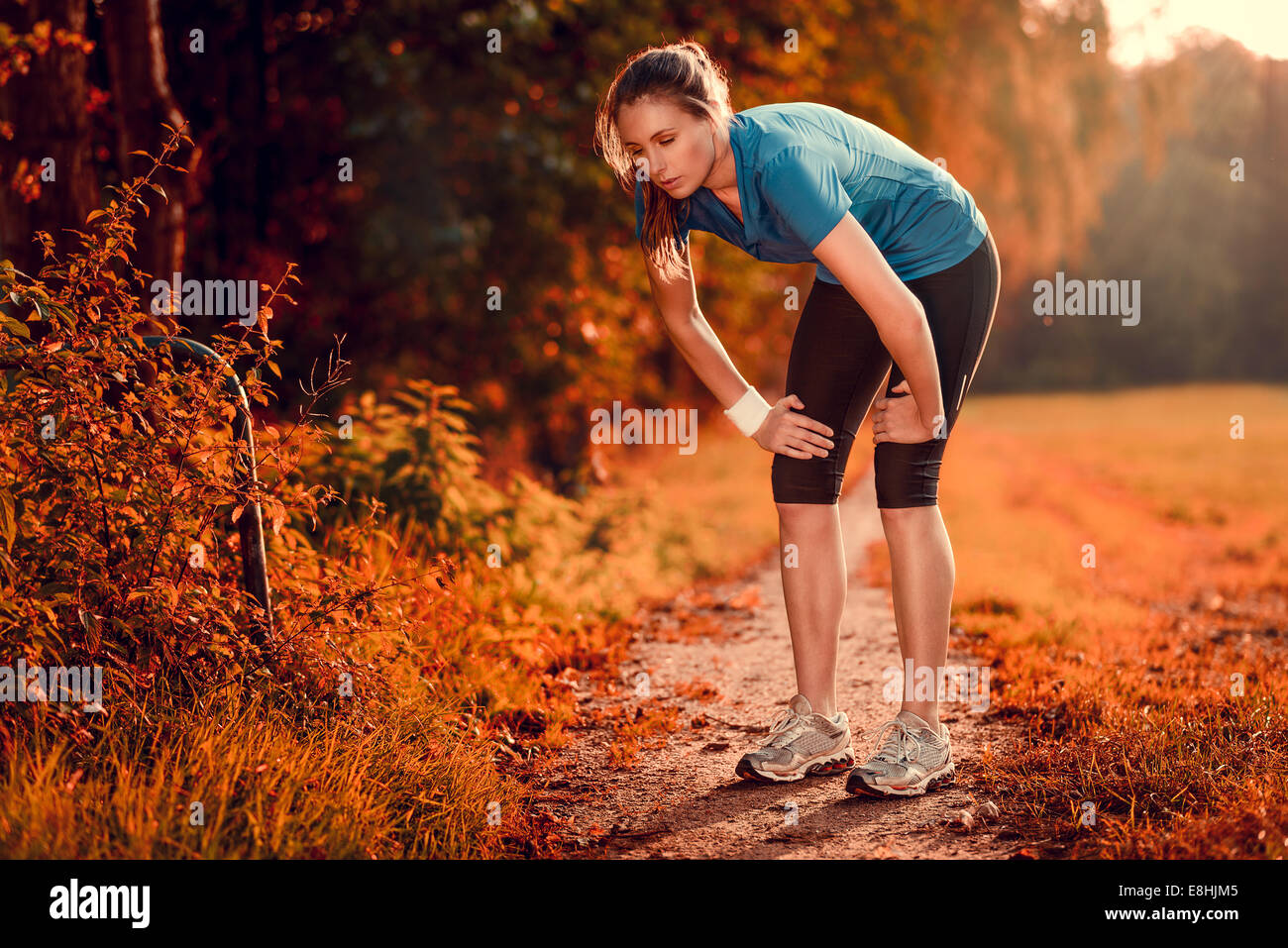 Junge sportliche Frau, die eine Pause vom Training stehen ruht ihr die Hände auf den Knien auf einem ländlichen Weg durch üppige Ackerland Stockfoto