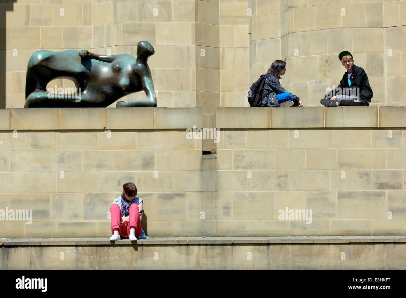 Leeds, UK. Menschen entspannen in der Sonne neben Henry Moore Skulptur "Liegende Frau" außerhalb der Leeds Art Gallery. Stockfoto