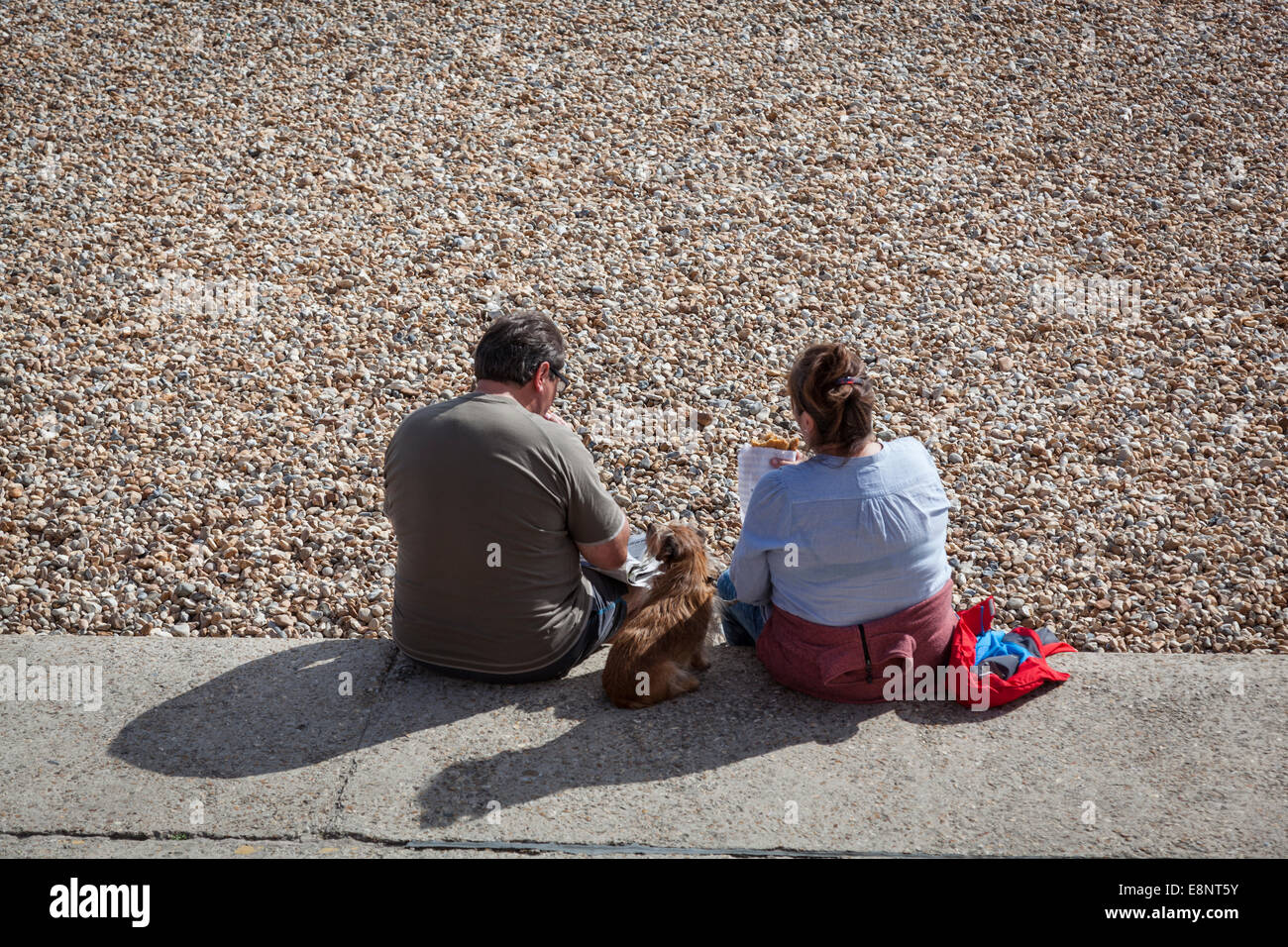 Paar, Fish &amp; Chips zu essen, am Rande von einem Kiesstrand mit einem Hund auf dazwischen sitzen. Stockfoto