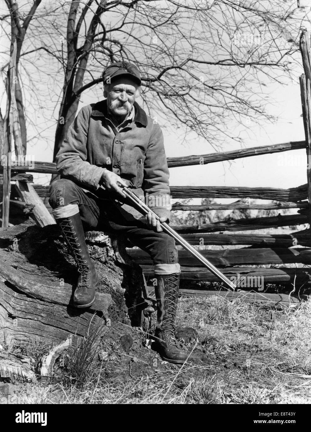 1940ER JAHRE ÄLTERER MANN SITZT AUF HOLZ SPLIT RAIL ZAUN HOLDING SCHUSS PISTOLE TRAGEN JAGDBEKLEIDUNG Stockfoto