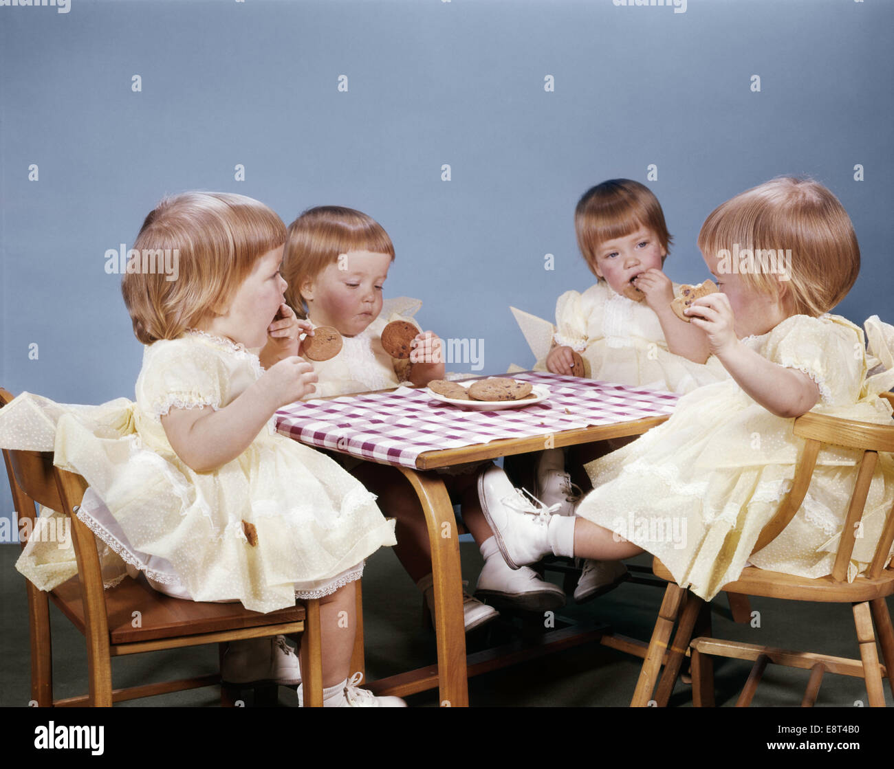 1960ER JAHREN VIERFACHER BABY MÄDCHEN SITZEN AM TISCH ESSEN COOKIES Stockfoto