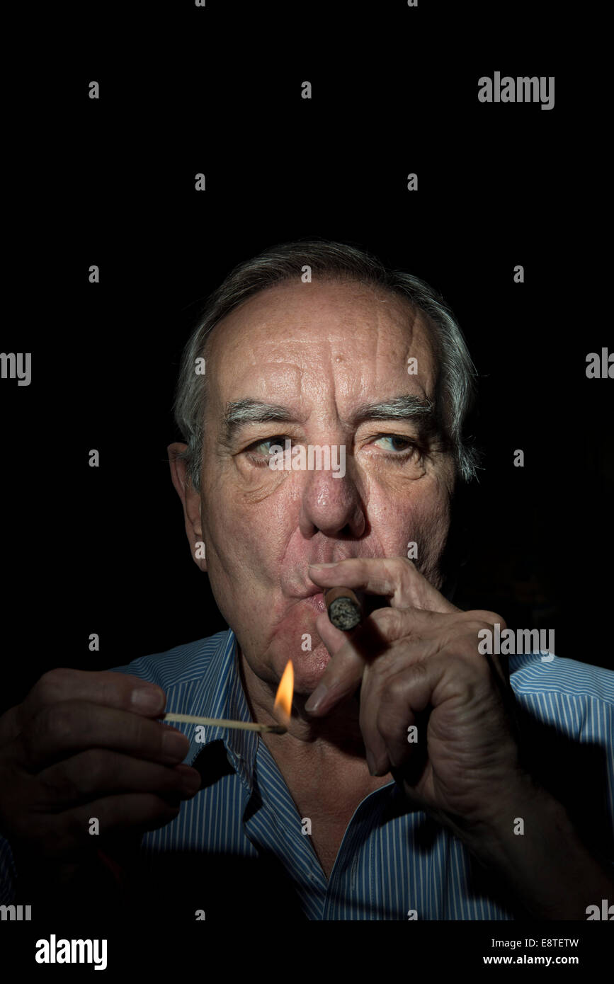 Ein Alter weißen Mann mit ein charaktervolles Gesicht leuchtet und raucht eine Zigarre vor einem schwarzen Hintergrund, mit einem dunklen & stimmungsvolle Foto Stockfoto