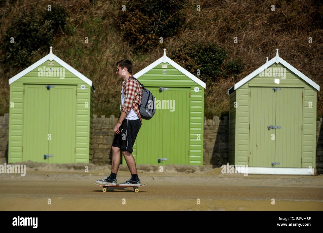 Ein Mann wird gesehen, skateboarding vorbei an einer Reihe von Strandhütten direkt am Strand in Bournemouth, England Stockfoto