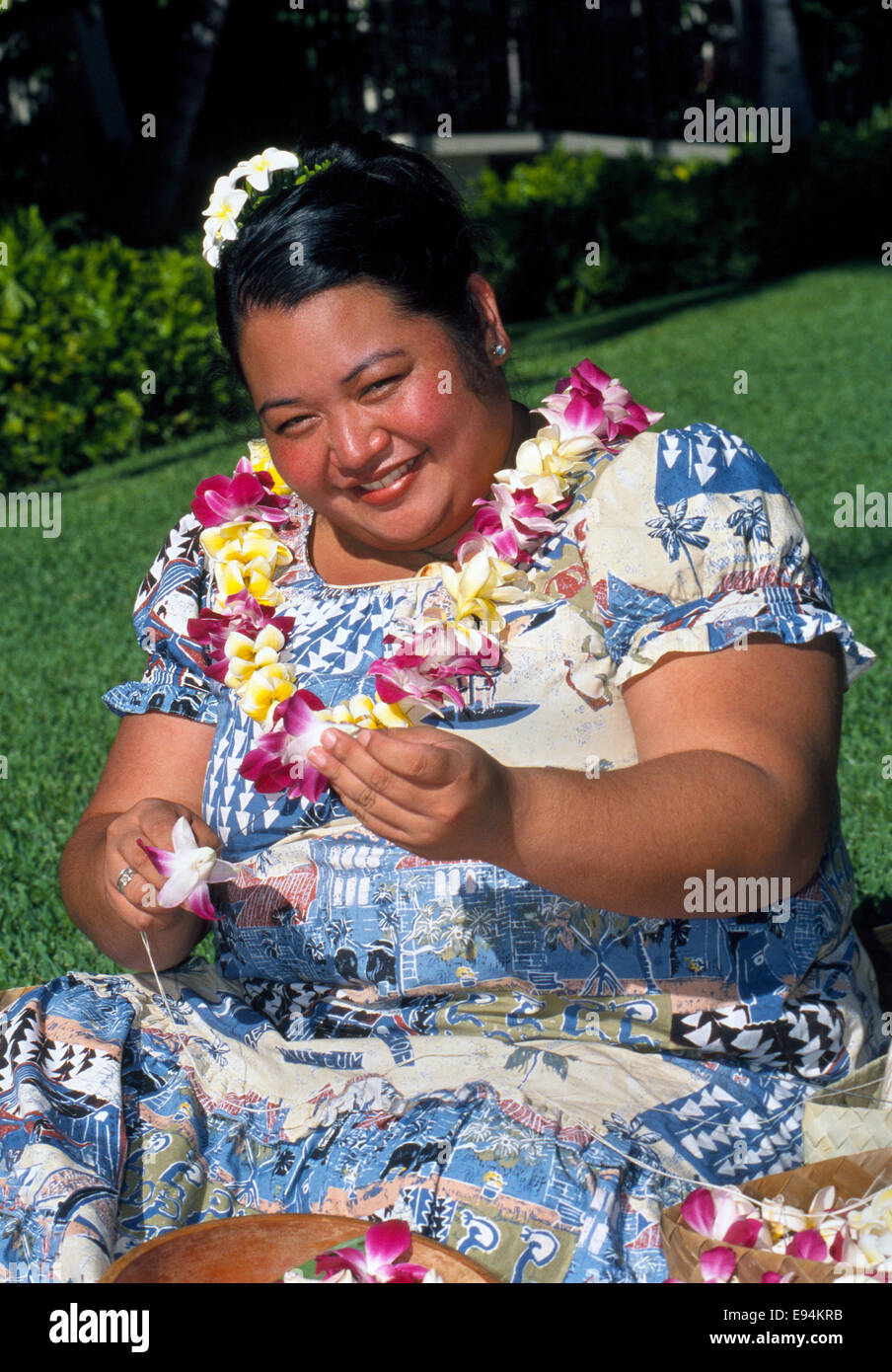 Eine lächelnde Frau Hawaii in Honolulu, Hawaii, USA, macht traditionellen Leis mit Plumeria (Frangipani) Blumen im Freien in der tropischen Sonne. Stockfoto