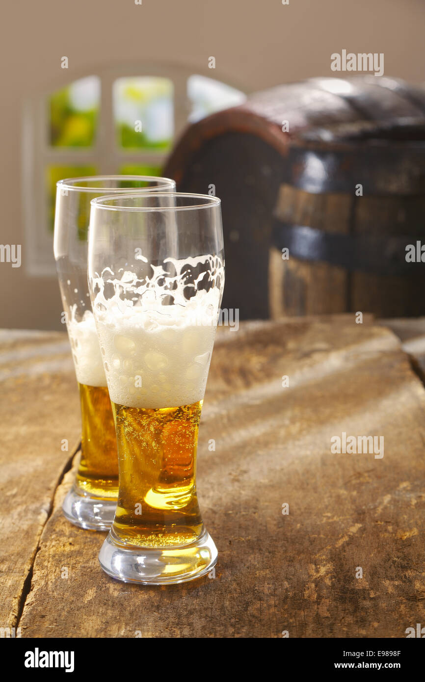 Zwei halb betrunken Gläser Bier mit einem guten schaumige Kopf stehen auf einem alten rustikalen Holztisch mit Vintage Holzfässern im Hintergrund Stockfoto