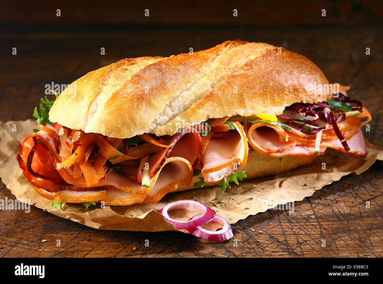 Leckere knusprige Baguette-Sandwich mit Schinken, Salat und Zwiebeln auf einem kleinen Grunge zerknittertes Blatt Packpapier auf einem Holztisch Stockfoto