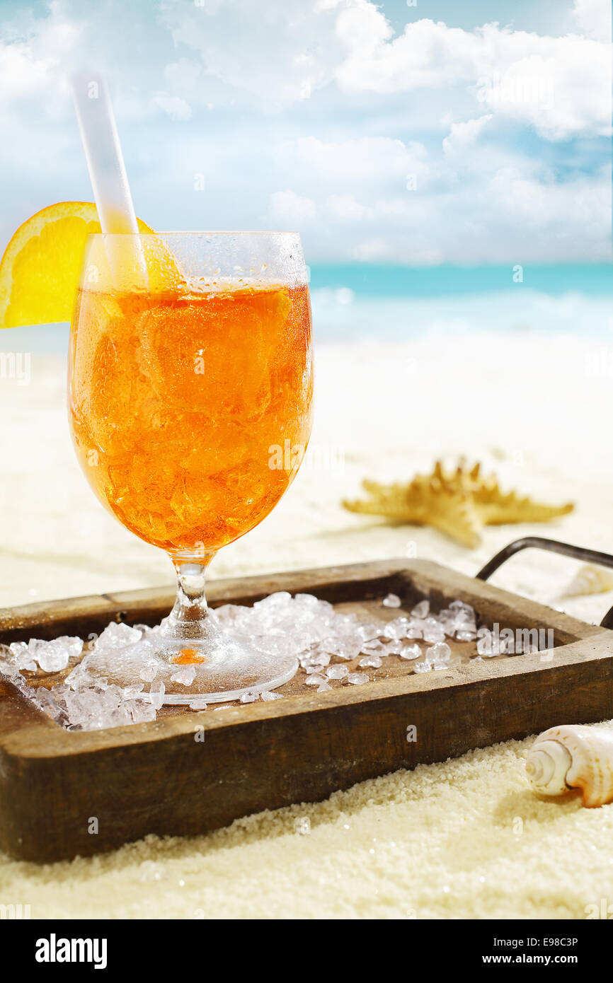 Leckeren eisgekühlten Aperol Spritzserved auf einem kleinen hölzernen Tablett verstreut mit Eiswürfeln auf einem heißen tropischen Strand im Sommersonnenschein Stockfoto