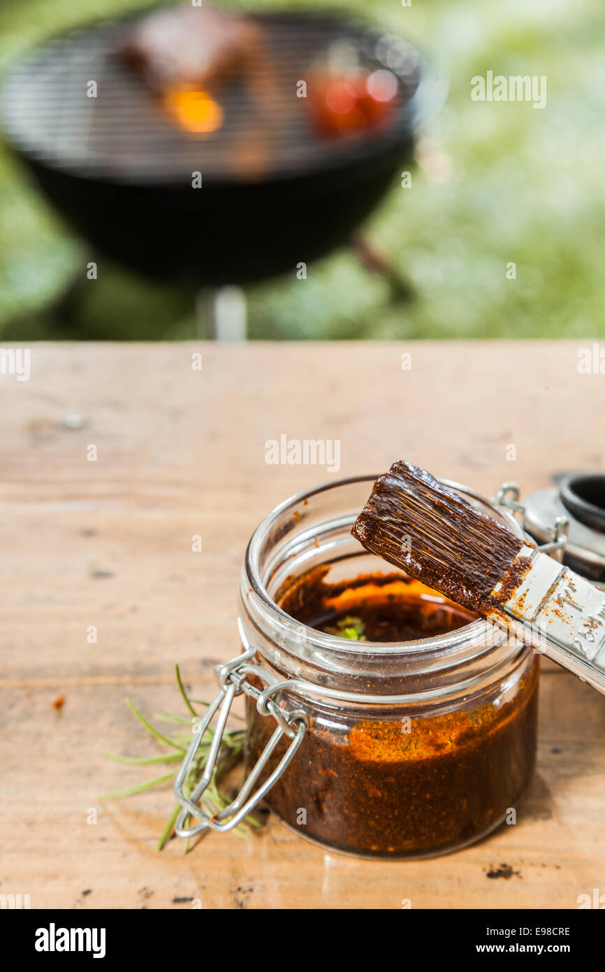 Glas mit Heften, Sauce und ein Heften Bürste stehend auf einem hölzernen Picknicktisch im Freien für das Fleisch auf dem Grill im Hintergrund auf einem grünen Rasen Grillen Stockfoto