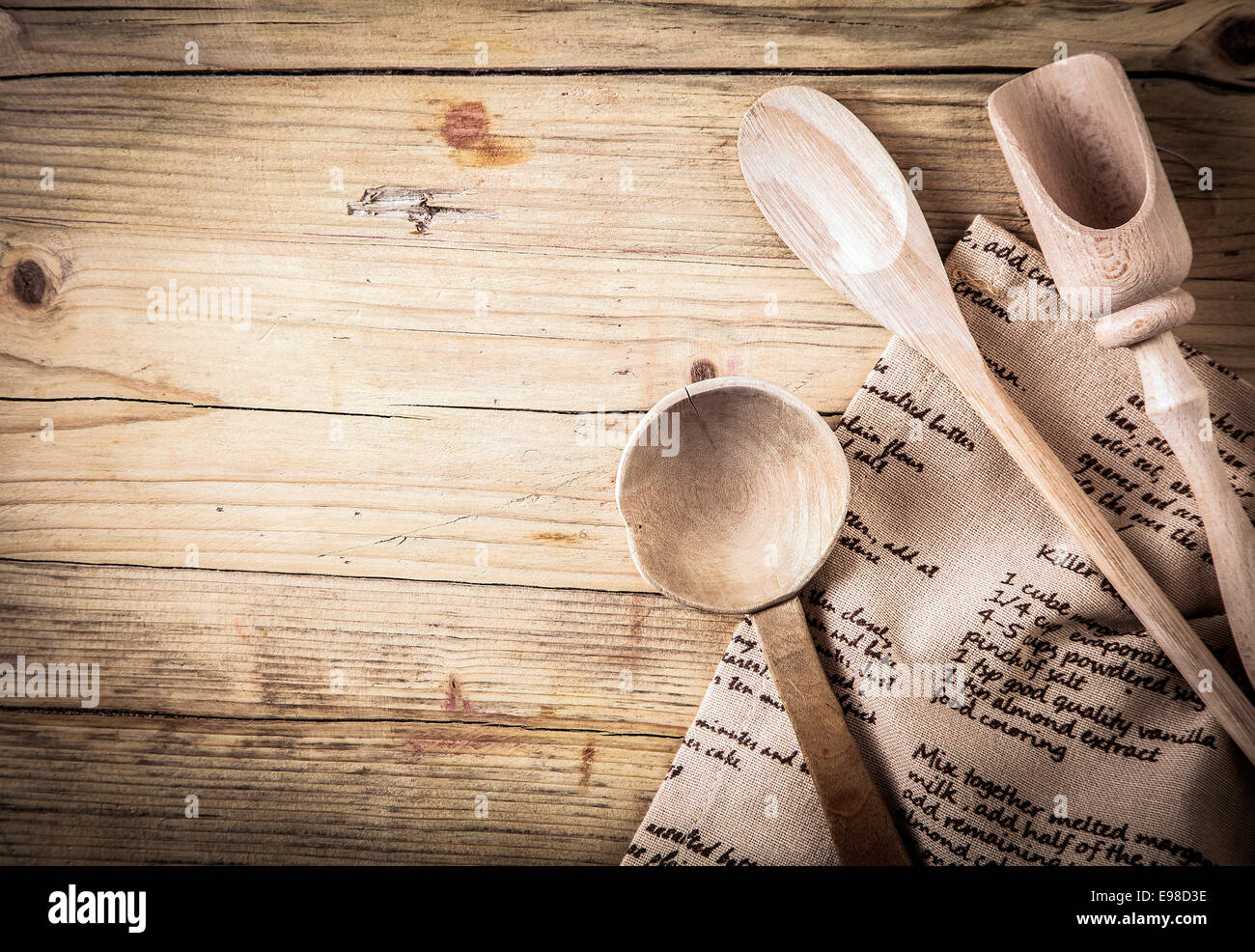 Rustikale Kochutensilien mit einem Holzlöffel, Kelle und Schaufel mit einem Rezept auf es auf einem alten gebrochenen Holztisch mit Vignettierung und Exemplar auf einem gefalteten Tuch liegend Stockfoto