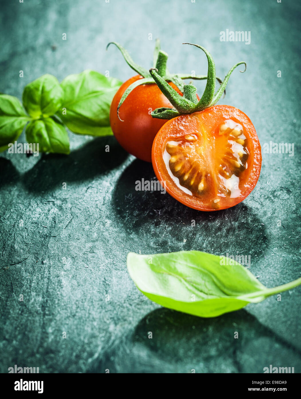 Frische Basilikumblätter mit halbierten saftige Reife rote Traube Tomaten bereit für das Kochen eines schmackhaften Gericht auf eine dunkle strukturierte Oberfläche Stockfoto