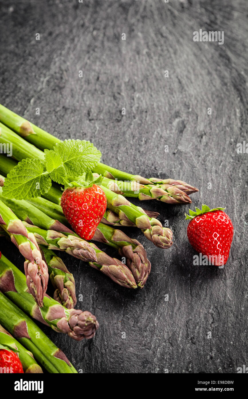 Frische ungekochte grünen Spargel, Erdbeeren und Pfefferminz auf einem grauen Schiefer Oberflächenstruktur mit Exemplar in Hochformat Stockfoto