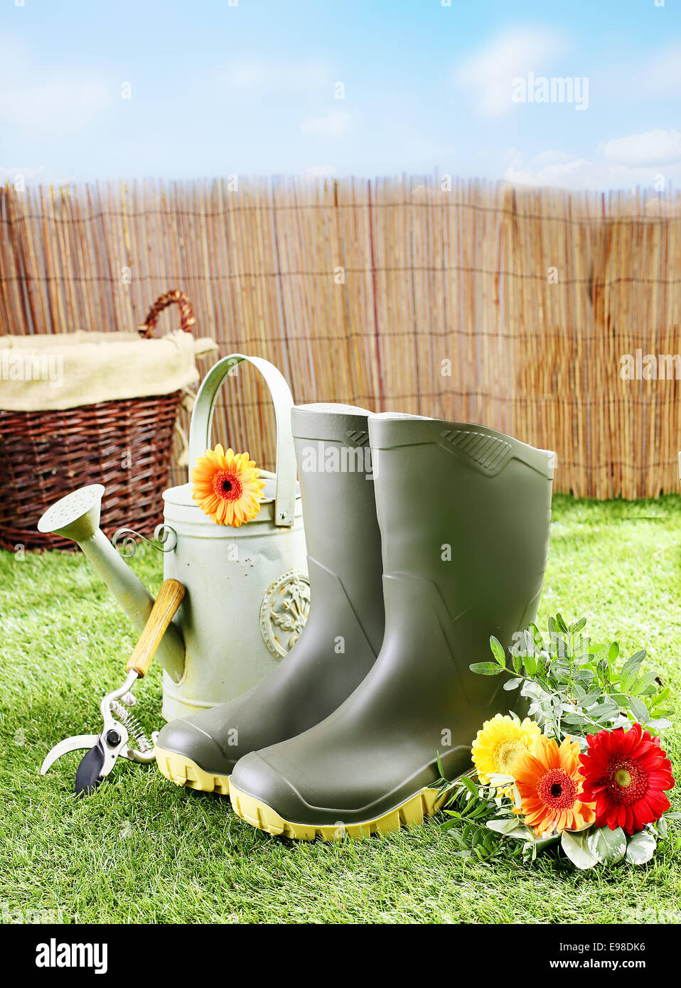 Gärtner-Werkzeuge und Geräte wie Gummistiefel, eine Gießkanne, Baum-, Rebscheren und einen Weidenkorb auf einem grünen Rasen und Blumen an einem sonnigen Tag Stockfoto