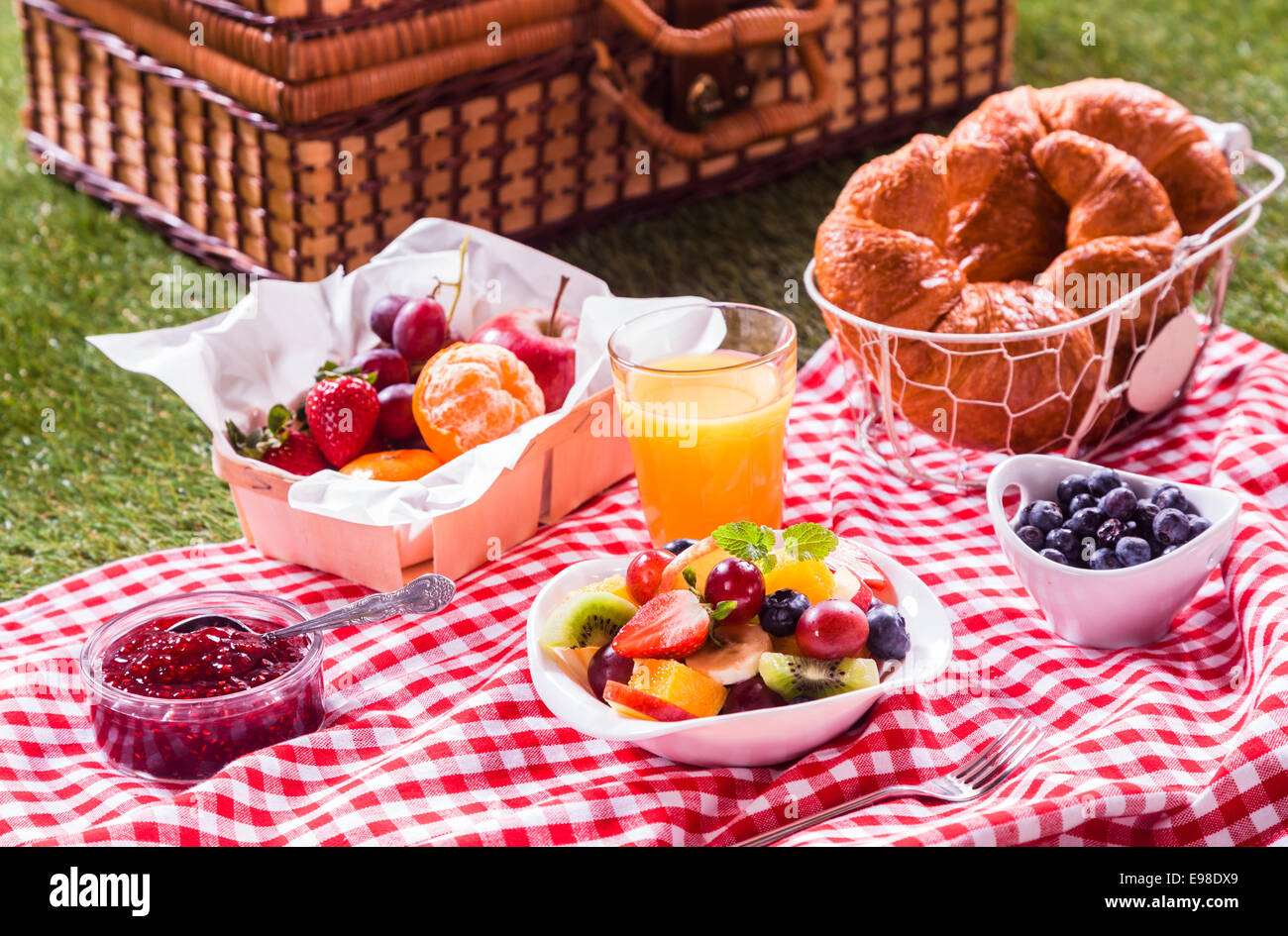 Gesunde vegetarische oder vegane Picknick mit einer köstlichen Auswahl an frisches Obst, goldene Croissants, Obstkonfitüre und tropischen Obstsalat auf eine rot-weiße Tischdecke neben einem Picknickkorb auf dem grünen Rasen Stockfoto