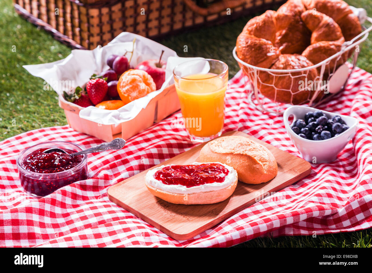 Köstliche Sommer-Picknick für Vegetarier ausgebreitet auf einem roten und weißen aufgegebenes Tuch auf dem Rasen mit goldenen frisch gebackenen Croissants, Orangensaft, frisches Obst und Obstsalat und ein Brötchen mit Marmelade Stockfoto