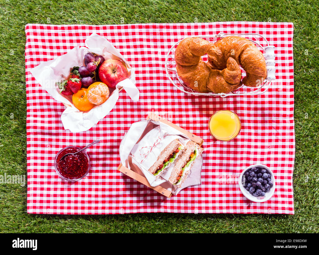 Genießen Sie eine gesunde Sommer-Picknick in der Sonne mit Sandwiches, Obst, Orangensaft, Marmelade, Croissants und Beere auf einem farbenfrohen roten und weißen Tuch auf einem grünen Rasen Stockfoto