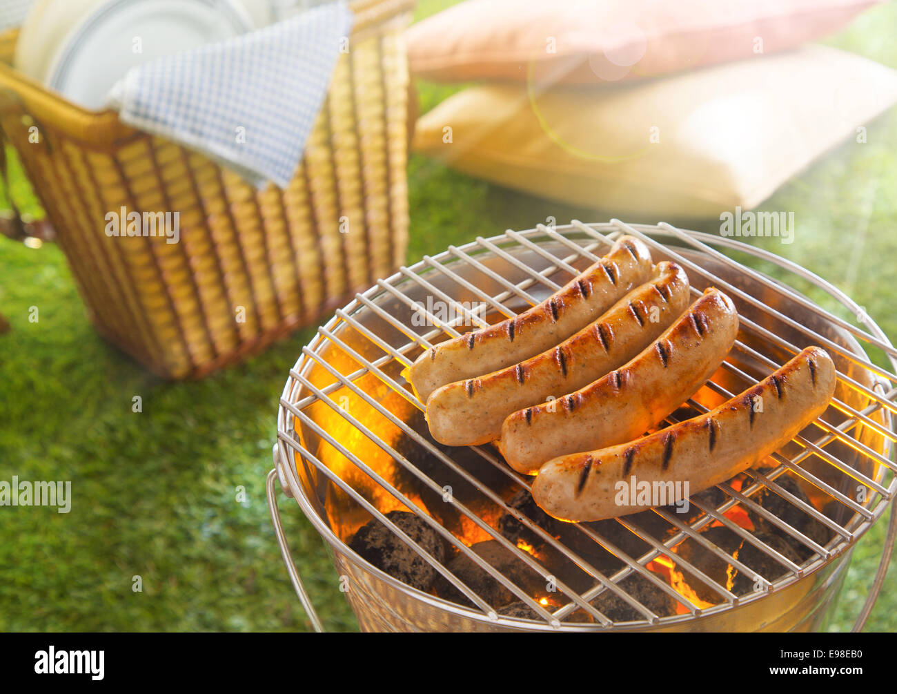Würstchen grillen oder glühend heißen Kohlen in einen tragbaren Metall Grill an einem Sommerpicknick mit einen geflochtenen Korb mit Platten und Kissen auf dem grünen Rasen neben Stockfoto