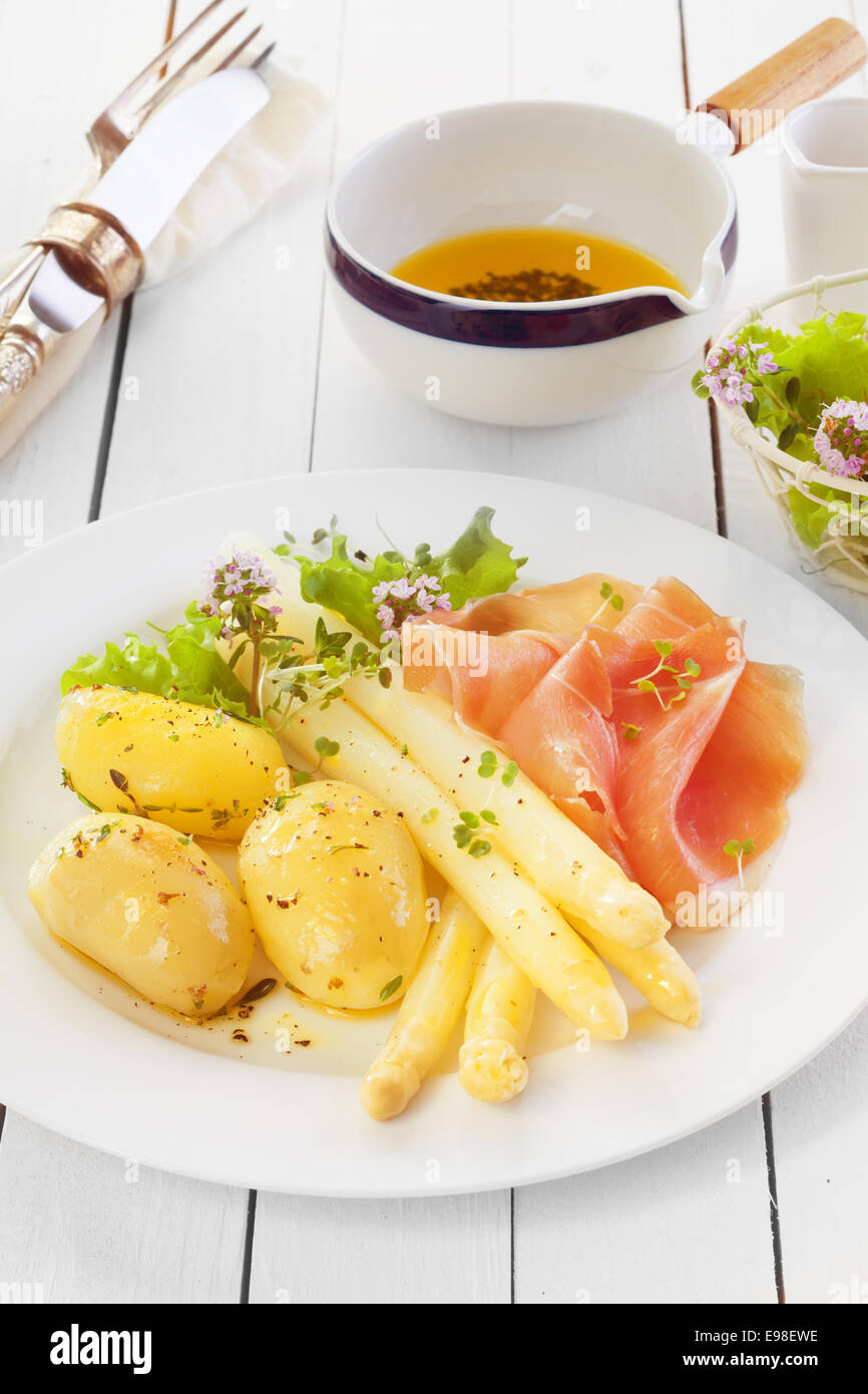 Gekochten Spargel mit italienischen Schinken und gekochte Babykartoffeln serviert mit Olivenöl und Kräuter dressing auf einer weißen hölzernen Theke in einer Landhausküche Stockfoto