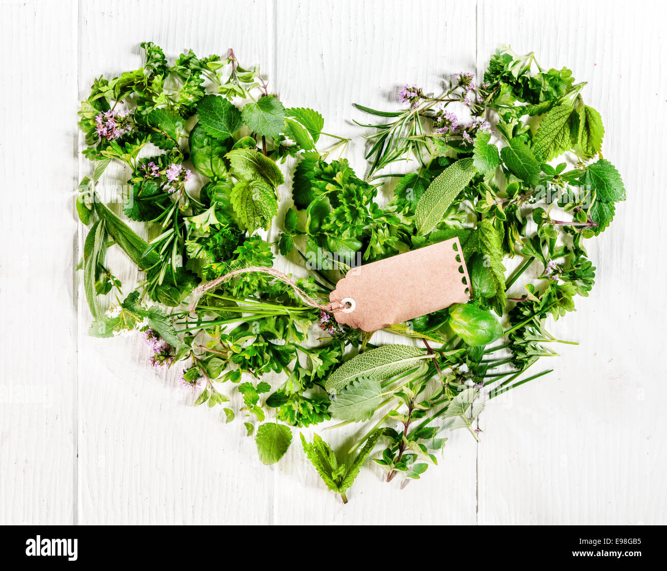 Herz, geformt aus sortierten frische grüne Küchenkräuter wie Petersilie, Thymian, Rosmarin, Basilikum und Schnittlauch mit einer leeren braun Geschenkanhänger auf rustikalen bemalten Tafeln symbolisch für Liebe und Romantik Stockfoto