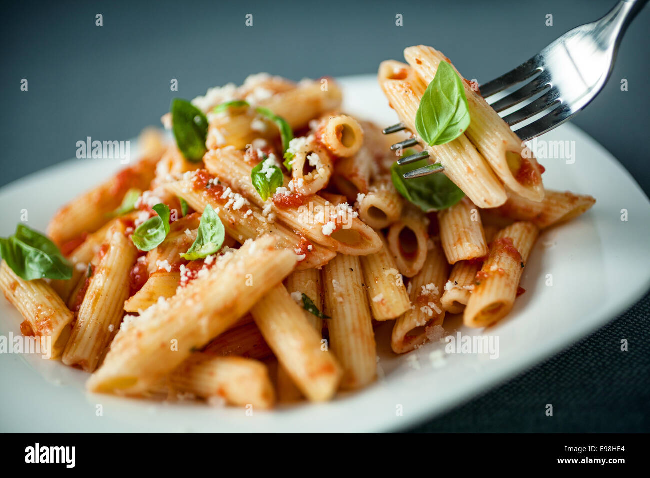 Penne-Nudeln mit pikanter Sauce, Basilikum und geriebenem Parmesan-Käse mit einer Gabel von Pasta über die Platte mit Fokus auf die Gabel aufgehängt Stockfoto