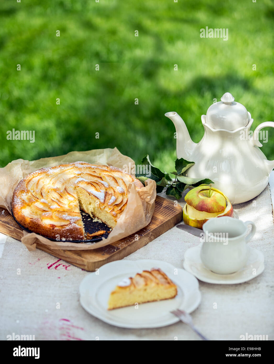 Kaffee oder Tee Pause im Garten mit einer frisch nach Hause gebackene Apfeltorte mit einer Scheibe serviert auf einem Teller mit eine leere Tasse und Untertasse und Tee oder Kaffee-Topf gegen grün Stockfoto