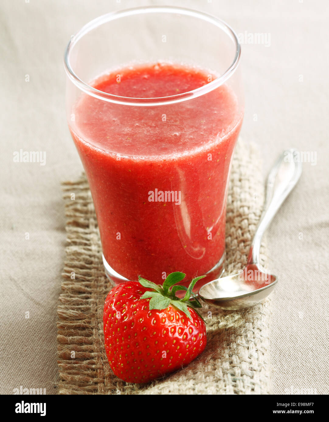 Glas frisch gemischt Erdbeer Smoothie auf Sackleinen mit einem üppigen Reife rote Erdbeere und silberne Löffel Stockfoto
