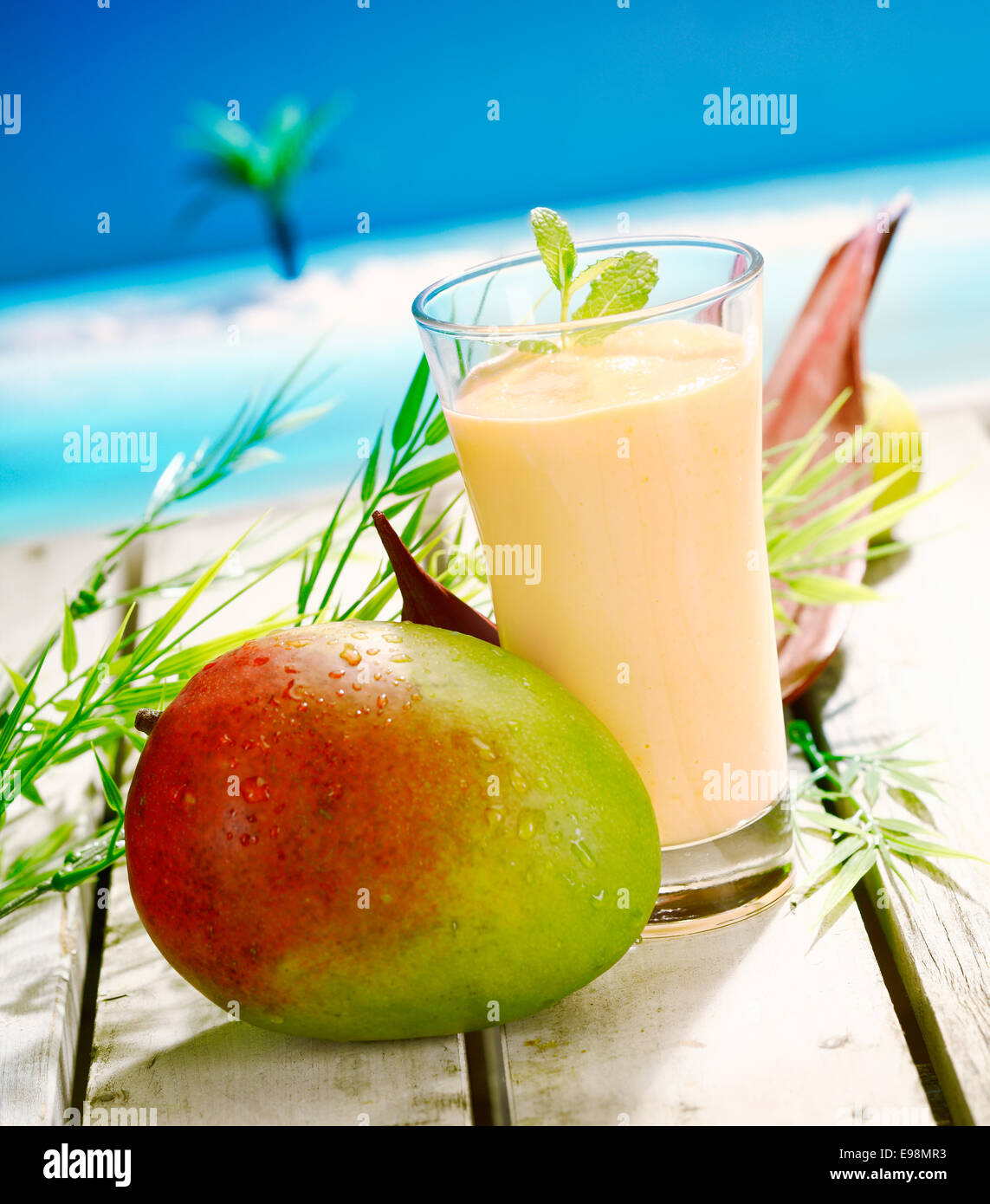 Glas von cremiger Mango und Joghurt Smoothie-Dessert mit eine große Reife Mango auf einem Holzdeck mit Blick aufs Meer Stockfoto