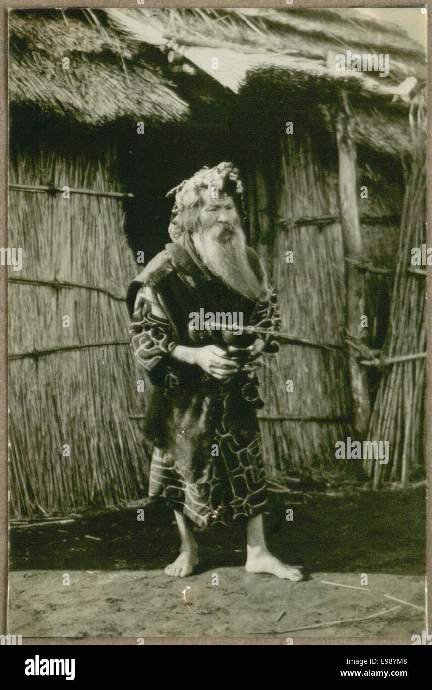 Dieses Foto zeigt eine Person namens Aino, die vor seinem Haus in Japan steht und Essen hält. Das Bild fängt einen Moment des Alltags im alten Japan ein und betont das traditionelle Haus und die traditionelle Kleidung. Stockfoto