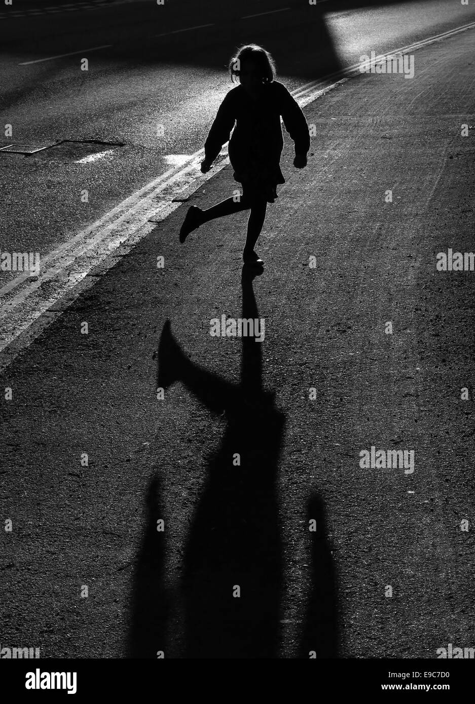 junge Mädchen am Straßenrand Silhouette durch auffallend helle niedrige Abendsonne auf einem Pflaster mit einem langen Schatten und Hintergrundbeleuchtung Haare tanzen Stockfoto