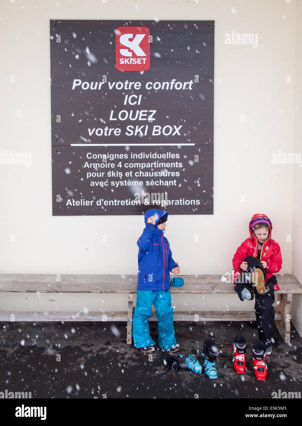 kleine Jungen und Mädchen in Skibekleidung sitzen auf einer Bank auf Skischuhe mit fallendem Schnee und ein Schild in Französisch hinter Stockfoto