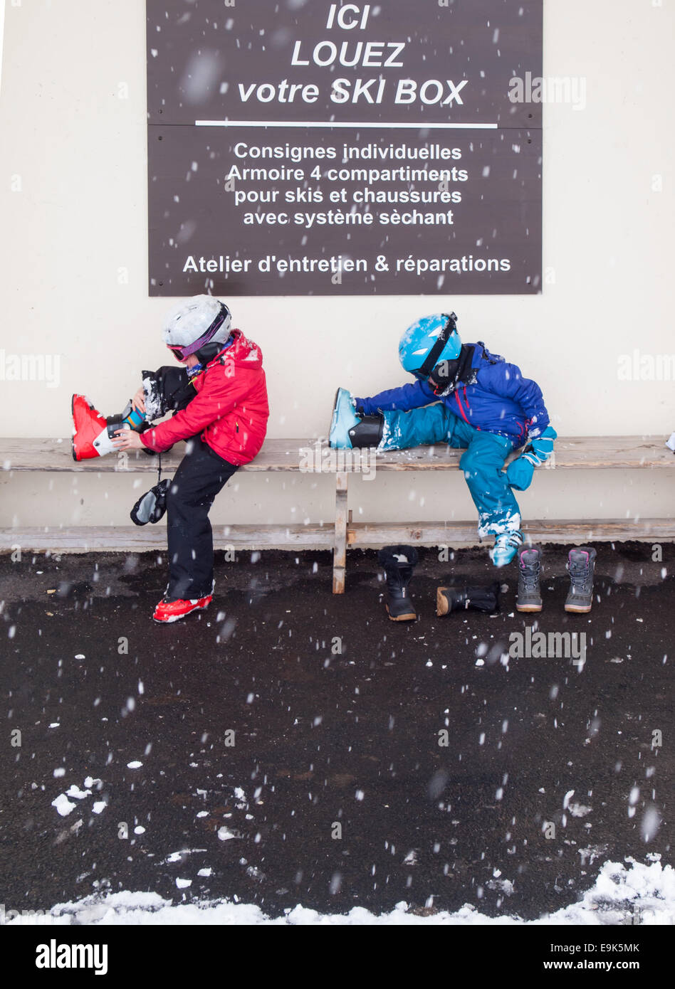 kleine Jungen und Mädchen in Ski-Bekleidung-Helme und Brillen sitzen auf einer Bank auf Skischuhe mit fallendem Schnee Stockfoto