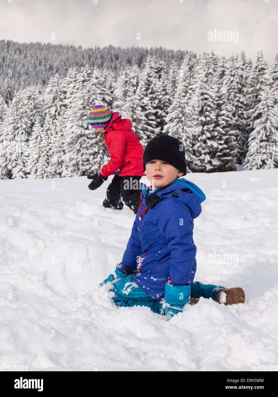 kleiner Junge spielt im Schnee mit kleinen Mädchen und bewaldeten Winter Berg Landschaft hinter Stockfoto