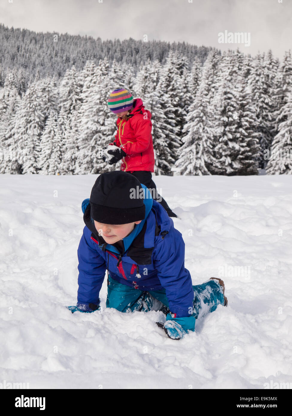kleiner Junge spielt im Schnee mit kleinen Mädchen und bewaldeten Winter Berg Landschaft hinter Stockfoto