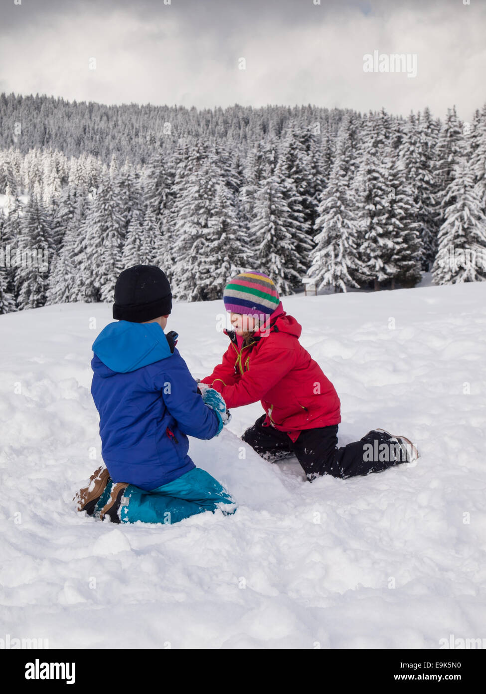 kleine Jungen und Mädchen spielen im Schnee mit bewaldeten Berglandschaft hinter winter Stockfoto