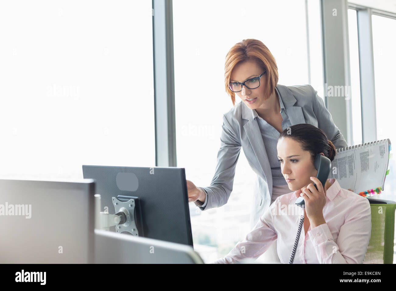 Junge Geschäftsfrau mit Festnetz-Telefon während Kollege zeigte auf Computer-Monitor im Büro Stockfoto