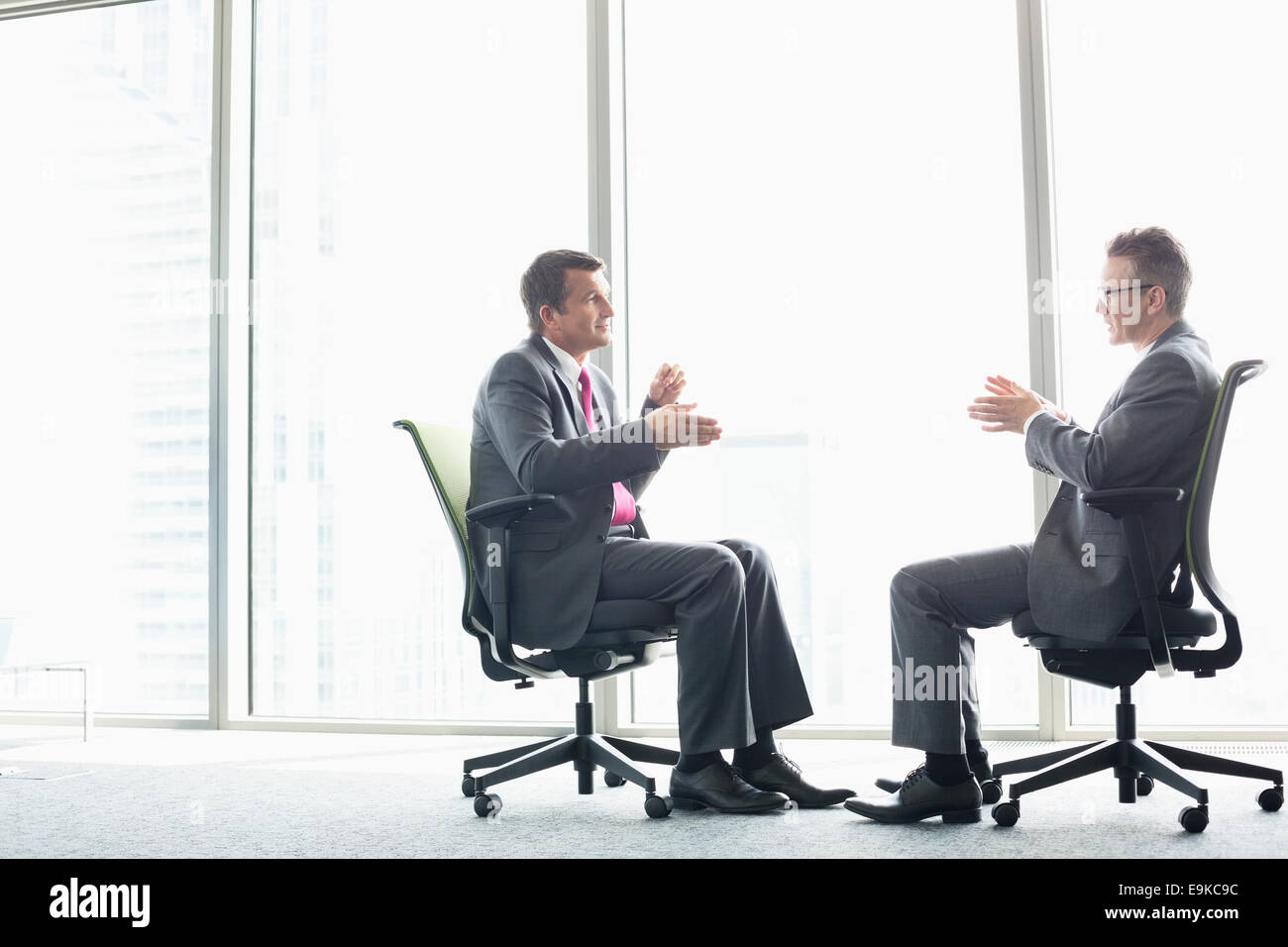 Full-length Seitenansicht der Unternehmer diskutieren beim Sitzen auf Bürostühlen durch Fenster Stockfoto