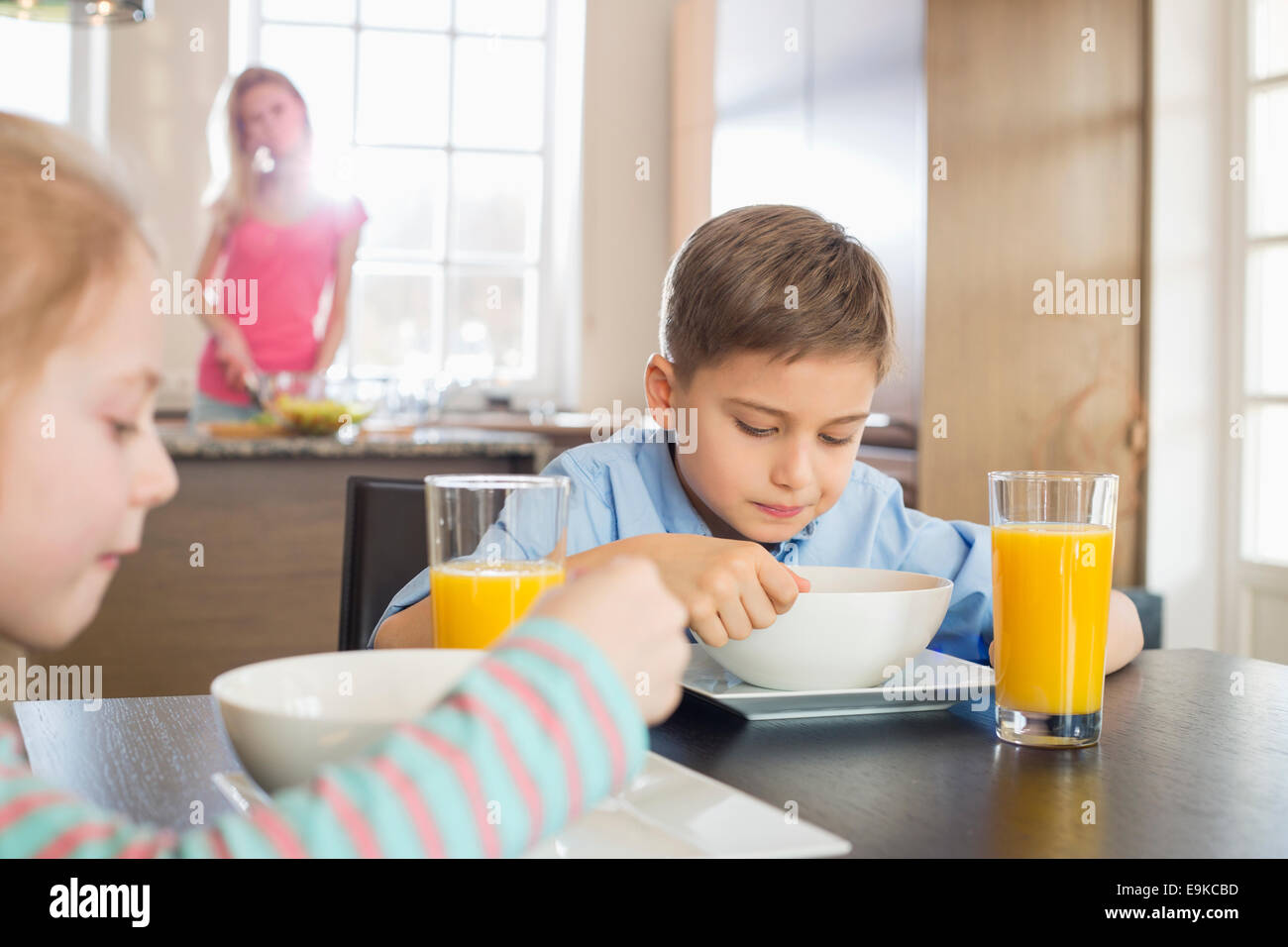 Geschwister mit Frühstück am Tisch mit Mutter, die Zubereitung von Speisen im Hintergrund Stockfoto