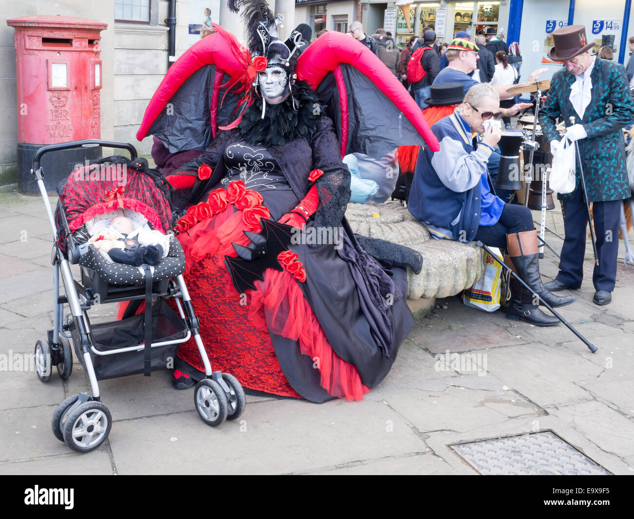 Eine Frau in einem aufblasbaren Fledermaus Kostüm sitzt mit einer Babypuppe gekleidet als Goten auf der Whitby Gothic Woche Ende Herbst 2014 Stockfoto