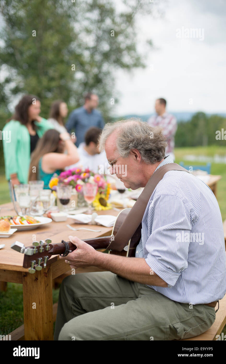 Reifer Mann, die Gitarre zu spielen, während die Freunde und Familie zusammen nach Essen im freien sprechen Stockfoto